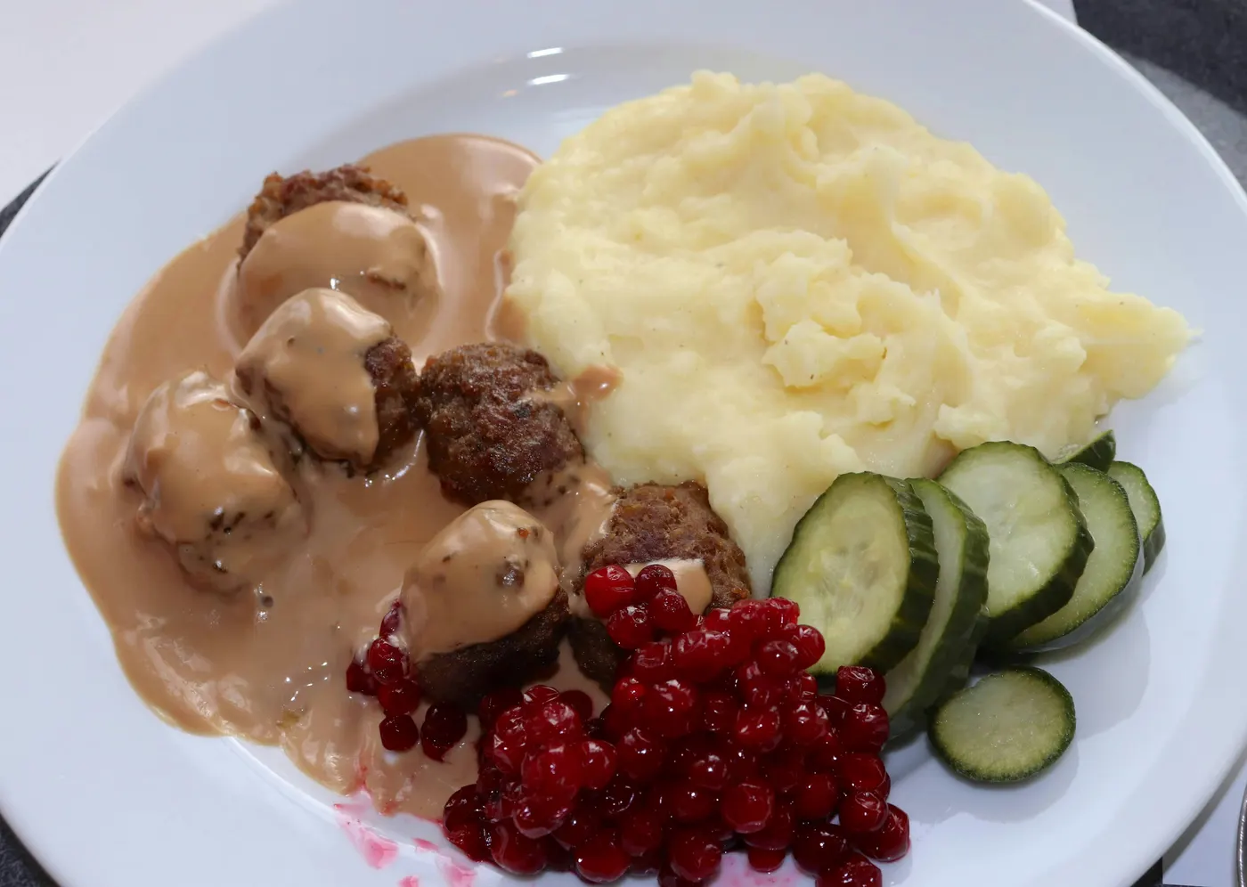Köttbullar med gräddsås, potatismos, lingon och pressgurka på en vit tallrik